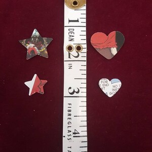 May include: Three paper cutouts of a star, a heart, and a star with a red, white, and blue design. The cutouts are placed on a red surface next to a white measuring tape with black markings. The tape measures in inches and is labeled "Dean" and "Fibreglass Tape".