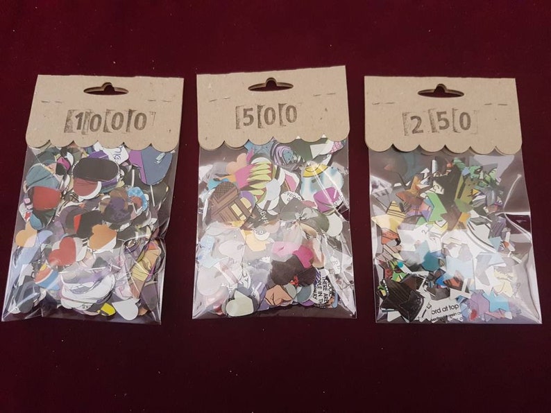 May include: Three bags of colourful paper confetti, each bag labelled with a different quantity: 100, 50, and 250. The confetti is made of various shapes and sizes, including hearts, stars, and circles.