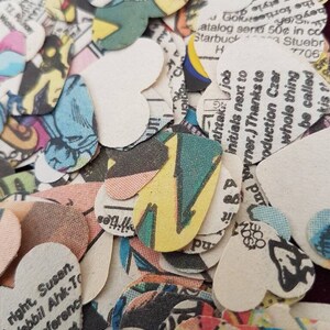 May include: A pile of heart-shaped confetti made from recycled comic book pages. The confetti is in various colors, including red, blue, green, yellow, and black. The confetti is printed with text and images from comic books.
