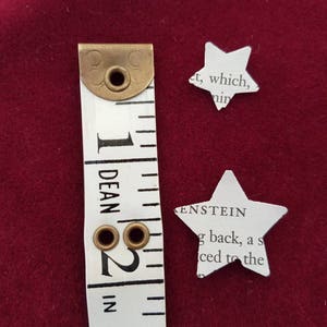 May include: A white measuring tape with brass eyelets and markings in inches. The tape is next to two white paper stars cut from a book. The text on the stars reads "et, which, nin" and "KENSTEIN g back, a s iced to the".