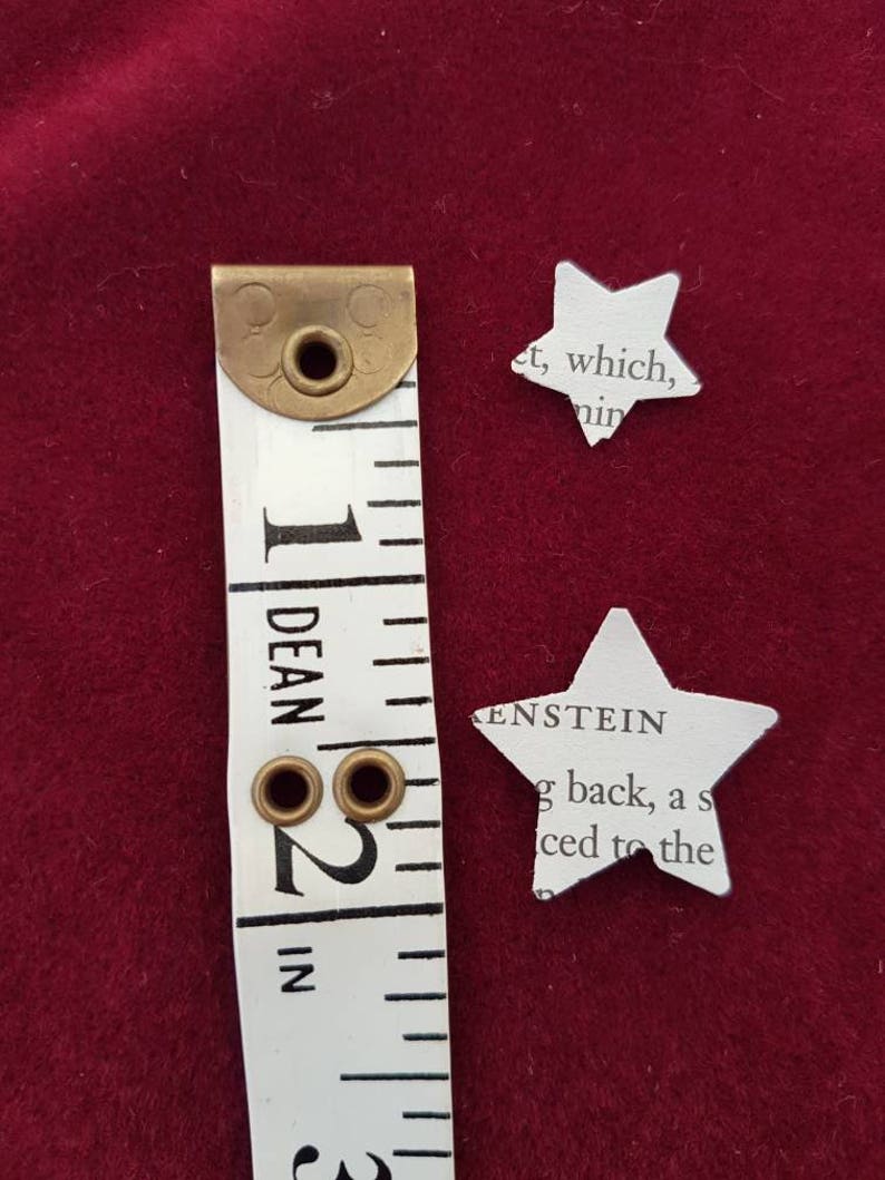 May include: A white measuring tape with brass eyelets and markings in inches. The tape is next to two white paper stars cut from a book. The text on the stars reads "et, which, nin" and "KENSTEIN g back, a s iced to the".