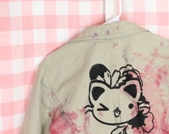 May include: Tan jacket with a black cat graphic on the back, accented with pink and purple paint splatters. The cat is winking and surrounded by floral elements. The jacket hangs on a white hanger against a pink and white checkered background.