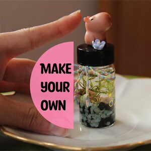 Könnte beinhalten: Ein kleines Glasflaschen-Terrarium mit schwarzem Deckel, gefüllt mit Miniaturpflanzen und Steinen. Auf dem Deckel sitzt eine kleine, hellbraune Capybara-Figur. Die Flasche ist mit farbigem Garn verziert und der Text "MAKE YOUR OWN" ist sichtbar.