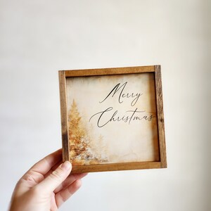 May include: A wooden framed sign with a watercolor background featuring a forest scene. The sign says "Merry Christmas" in black cursive lettering.