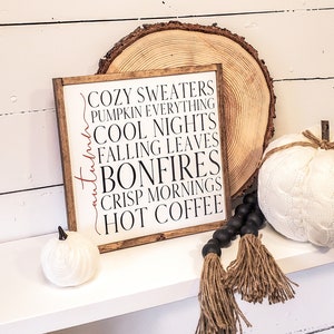May include: A wooden sign with the text "Cozy Sweaters, Pumpkin Everything, Cool Nights, Falling Leaves, Bonfires, Crisp Mornings, Hot Coffee" in black lettering with the word "Autumn" in red lettering. The sign is on a white shelf with a white pumpkin and a black and brown tassel garland.