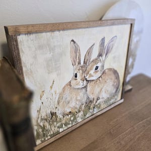 May include: A framed painting depicting two brown rabbits with long ears, snuggled together in a field of green grass and wildflowers. The painting has a rustic, vintage aesthetic.