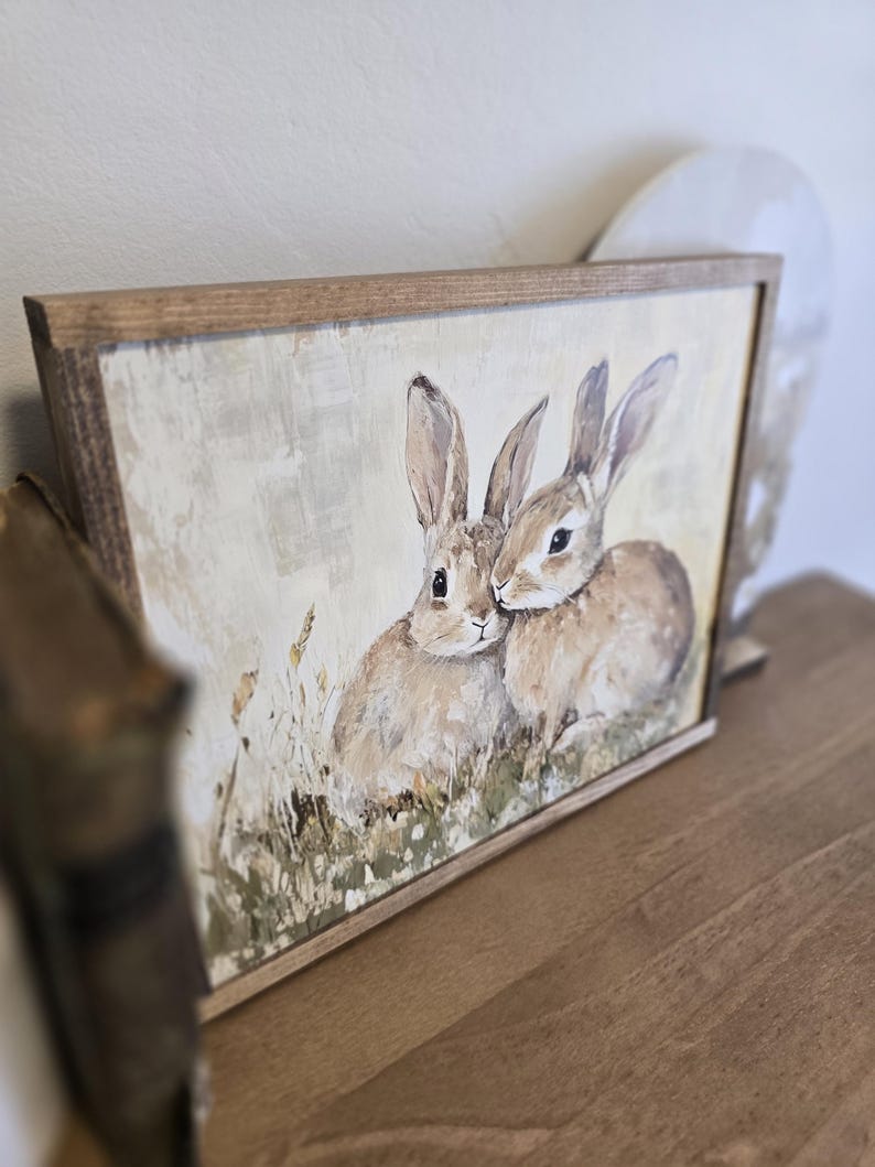 May include: A framed painting depicting two brown rabbits with long ears, snuggled together in a field of green grass and wildflowers. The painting has a rustic, vintage aesthetic.