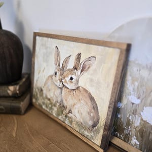 May include: A framed painting of two brown rabbits with long ears, sitting in a field of green grass. The painting has a light yellow background and is framed in a rustic brown wood frame.