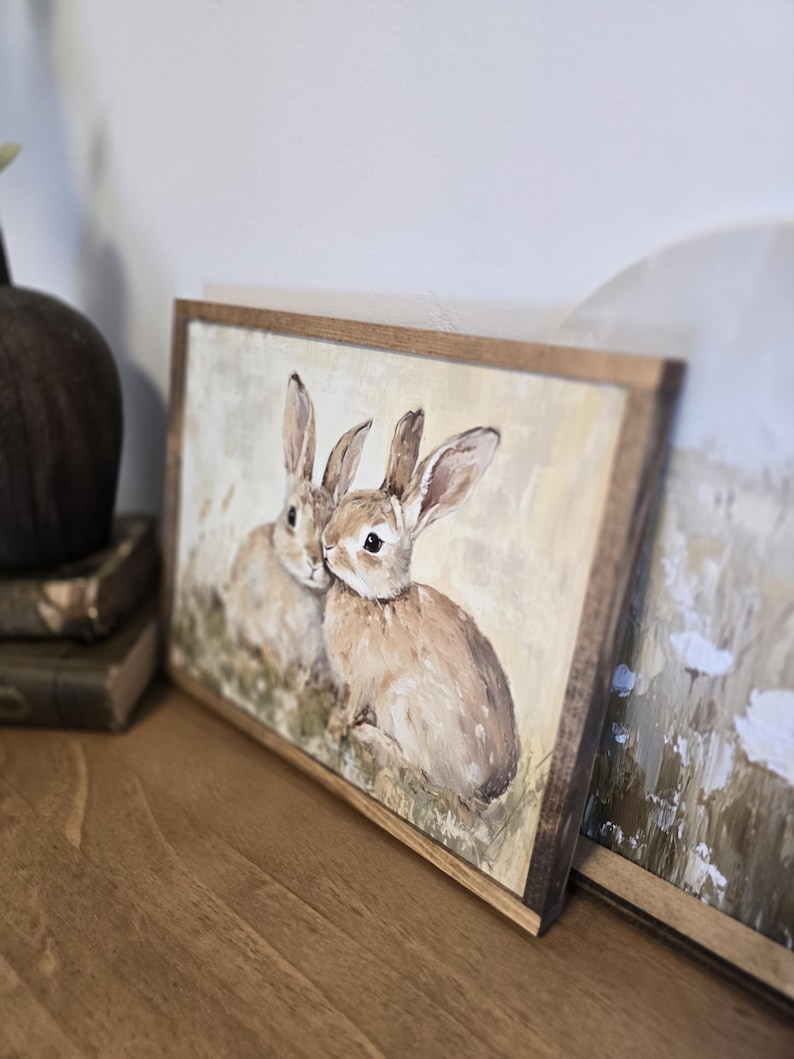 May include: A framed painting of two brown rabbits with long ears, sitting in a field of green grass. The painting has a light yellow background and is framed in a rustic brown wood frame.