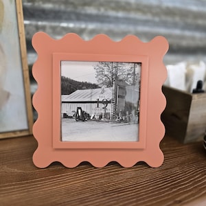 May include: A square, peach-colored picture frame with a scalloped edge. The frame holds a black and white photograph of a building with the text "THE 19 COLLECTIVE" visible. The frame is displayed on a wooden surface.