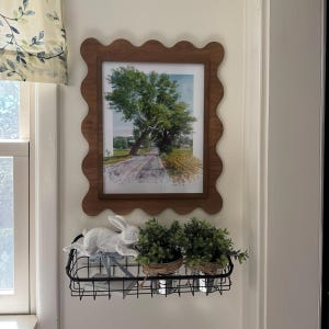 May include: A framed landscape print featuring a tree on a road, displayed on a wall. The frame is a wavy-edged wood. Below, a black wire basket holds a white rabbit figurine and two small potted plants.