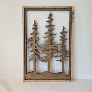 May include: A wooden wall decor featuring a silhouette of three pine trees in a brown frame.
