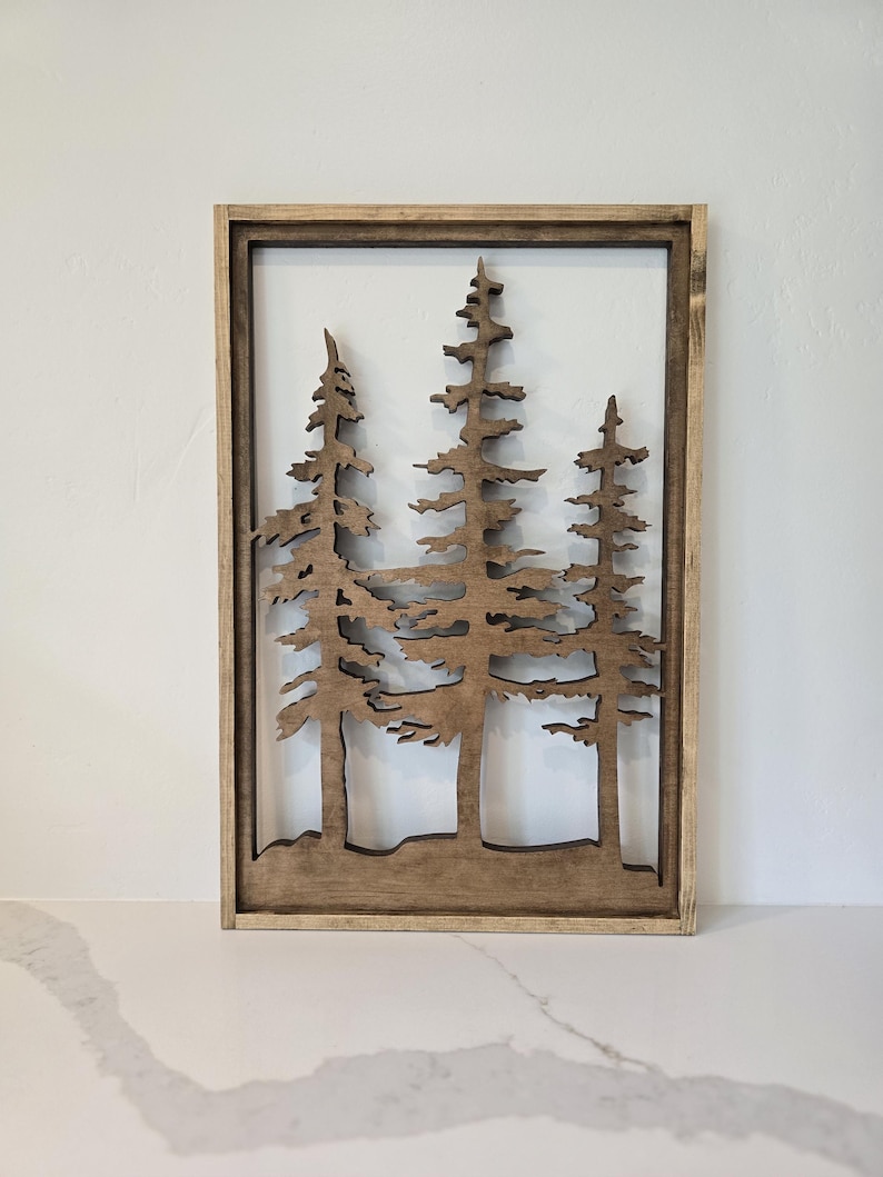 May include: A wooden wall decor featuring a silhouette of three pine trees in a brown frame.