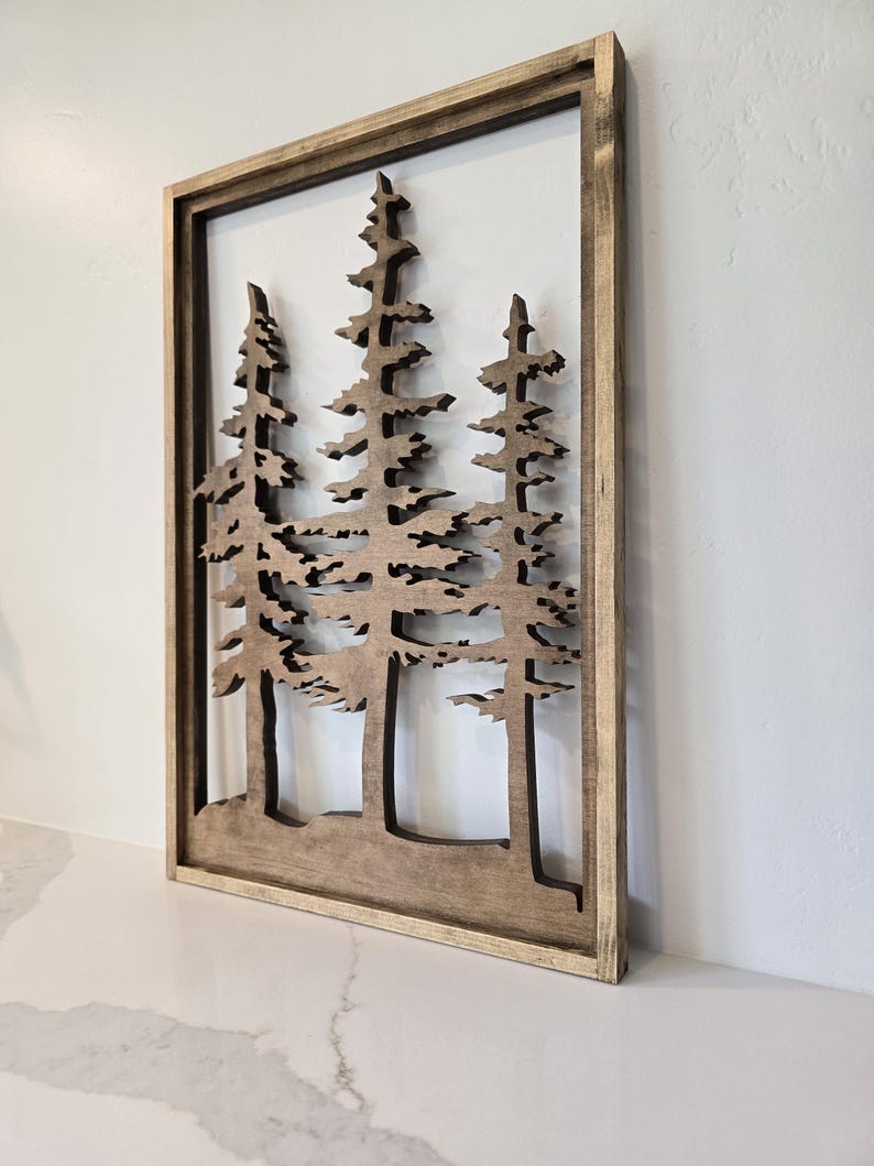 May include: A wooden wall decor featuring a cutout design of three pine trees. The trees are brown and have a rustic look. The decor is framed in a natural wood frame.