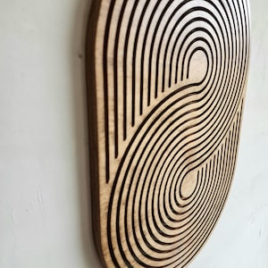 May include: A wooden wall decor with a circular design featuring concentric circles and curved lines. The design is reminiscent of a stylized eye or a swirling galaxy.