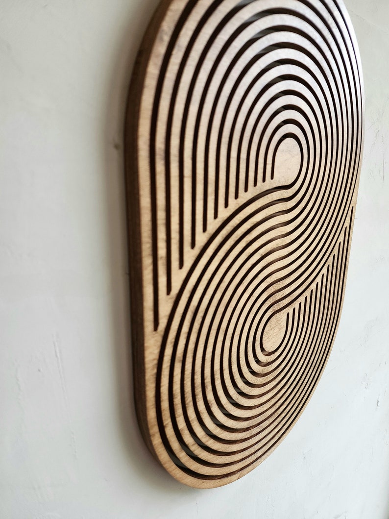 May include: A wooden wall decor with a circular design featuring concentric circles and curved lines. The design is reminiscent of a stylized eye or a swirling galaxy.