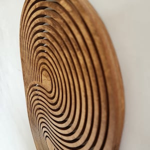 May include: A wooden wall decor with a circular design featuring concentric circles and a wavy pattern. The wood is a light brown color.