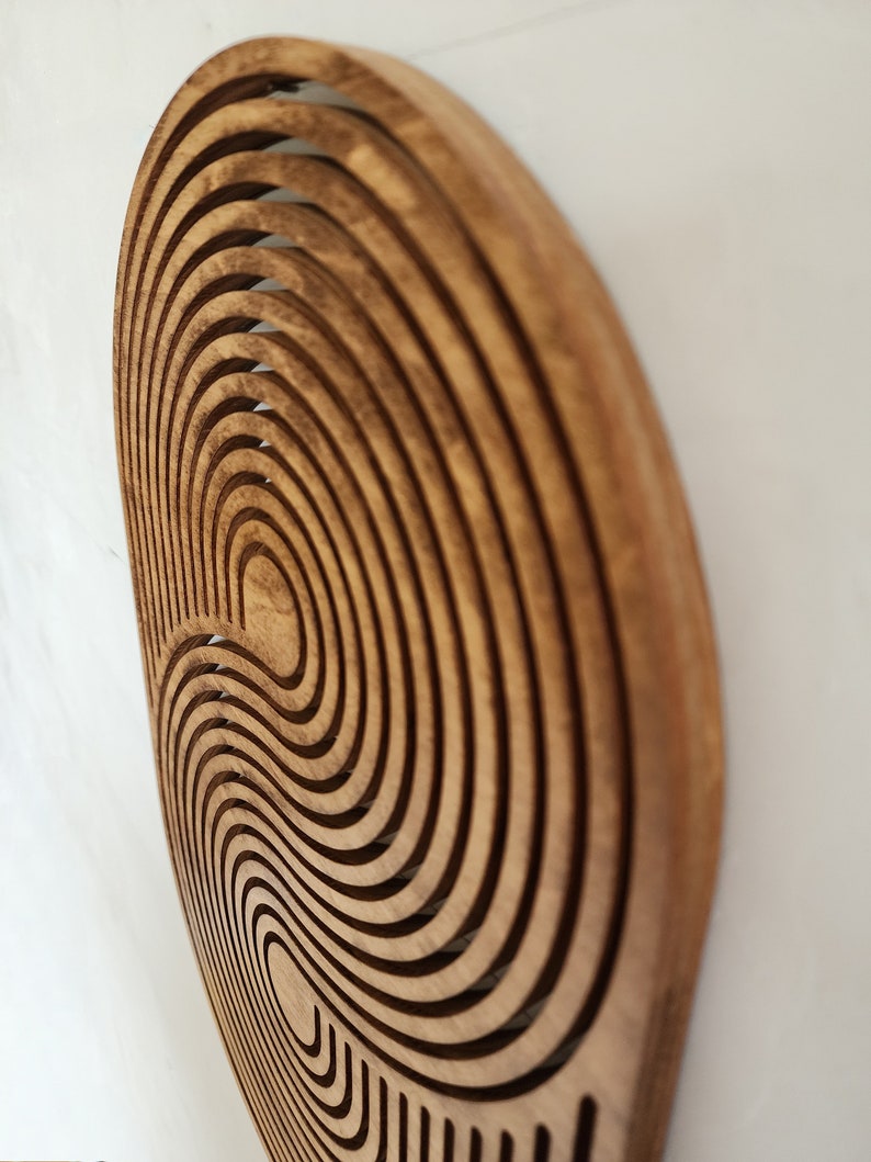May include: A wooden wall decor with a circular design featuring concentric circles and a wavy pattern. The wood is a light brown color.