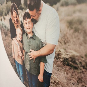 May include: A family of four poses for a photo in a field. The father is kissing his son on the head, while the mother and younger son stand beside them. The family is wearing casual clothing and smiling.