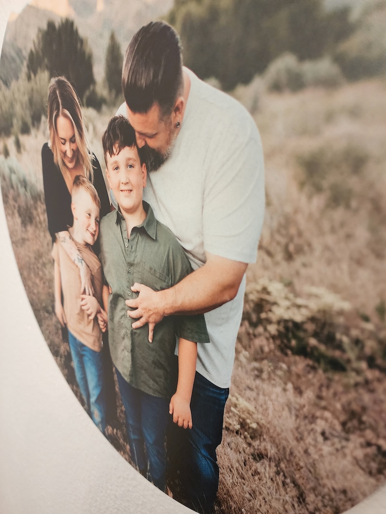 May include: A family of four poses for a photo in a field. The father is kissing his son on the head, while the mother and younger son stand beside them. The family is wearing casual clothing and smiling.