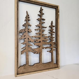 May include: A wooden wall art piece featuring a silhouette of three pine trees in a brown frame. The trees are cut out of the wood and the background is a light brown color.