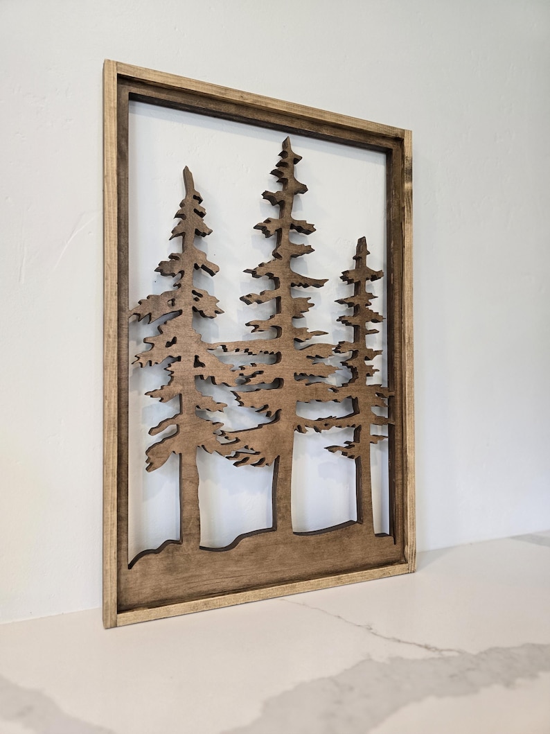 May include: A wooden wall art piece featuring a silhouette of three pine trees in a brown frame. The trees are cut out of the wood and the background is a light brown color.