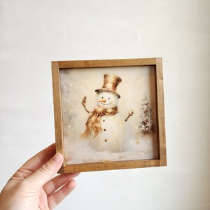 May include: A framed illustration of a snowman with a brown top hat and scarf, standing in a snowy landscape. The frame is made of wood and has a rustic finish.