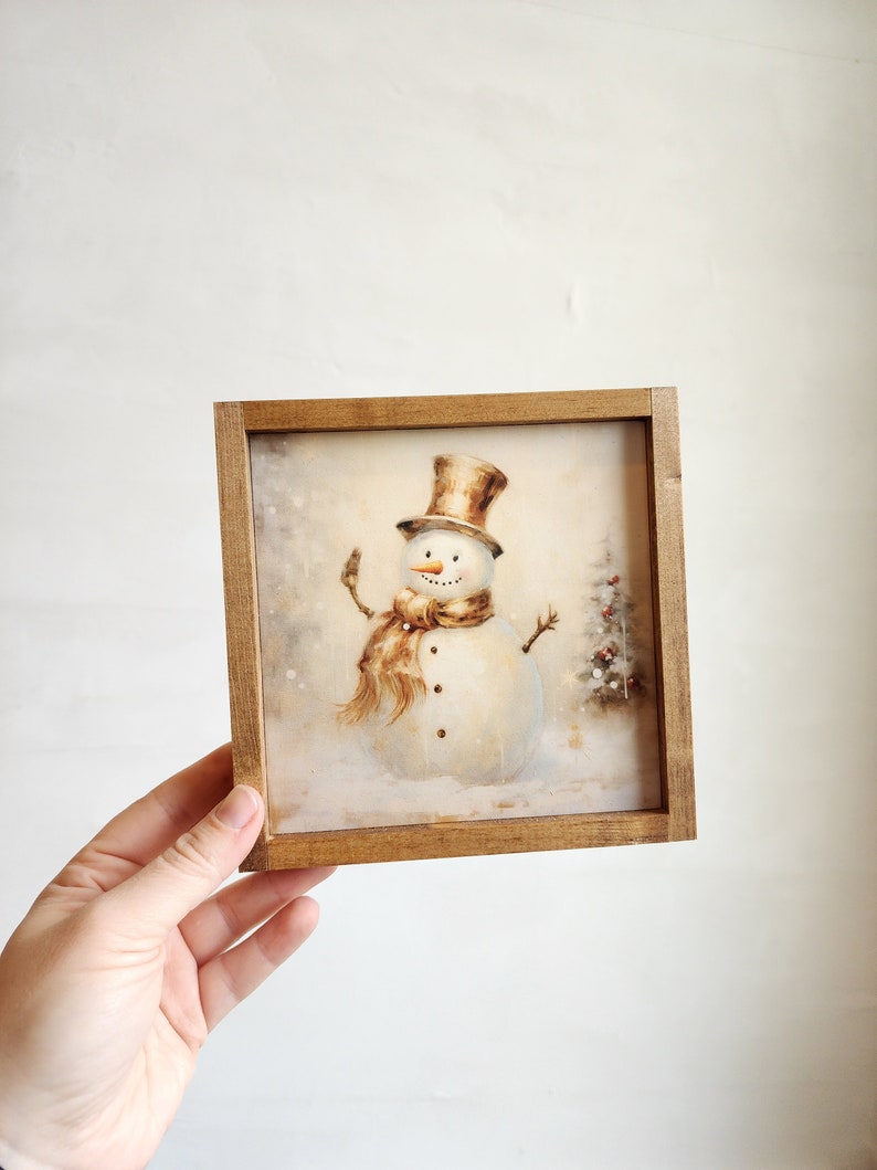 May include: A framed illustration of a snowman with a brown top hat and scarf, standing in a snowy landscape. The frame is made of wood and has a rustic finish.