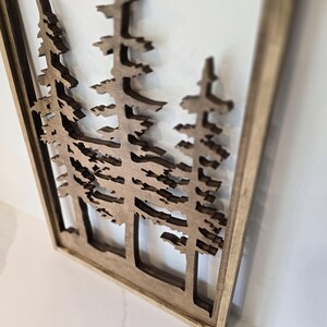 May include: A wooden wall art piece featuring a cutout design of three pine trees. The trees are brown and the background is white. The piece is framed in a natural wood frame.