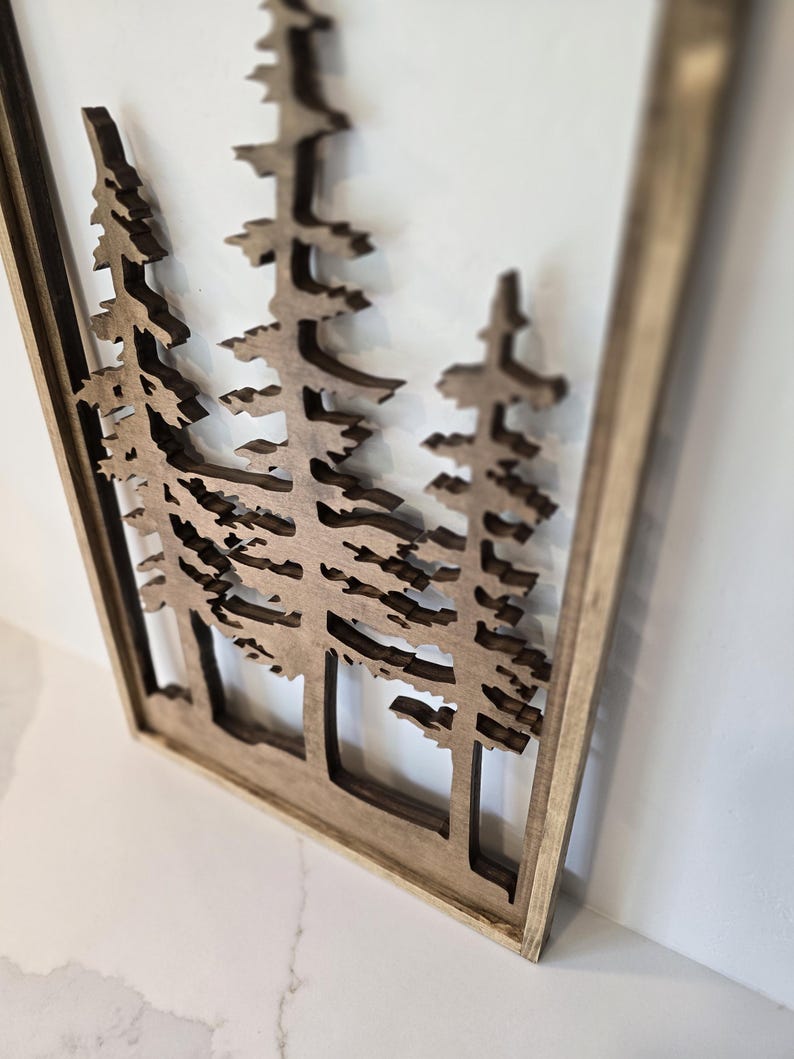 May include: A wooden wall art piece featuring a cutout design of three pine trees. The trees are brown and the background is white. The piece is framed in a natural wood frame.
