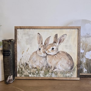 May include: A framed painting depicting two brown rabbits cuddling in a field of tall grass. The painting has a light beige background with a soft, painterly style.