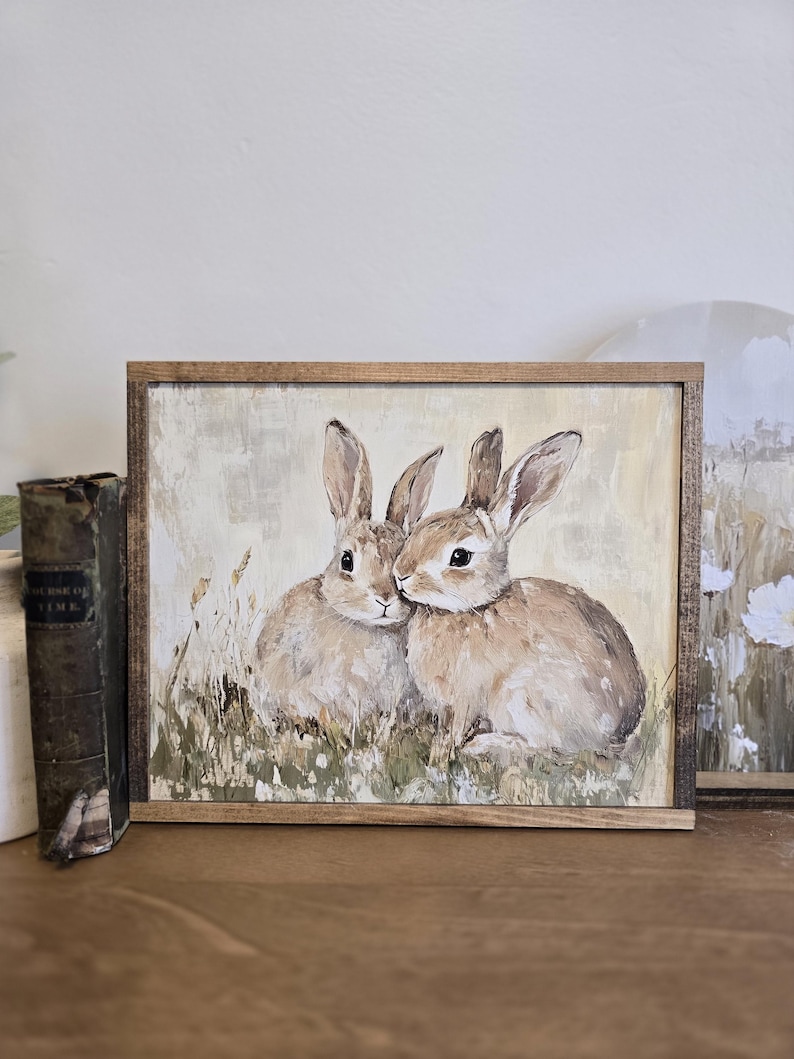 May include: A framed painting depicting two brown rabbits cuddling in a field of tall grass. The painting has a light beige background with a soft, painterly style.