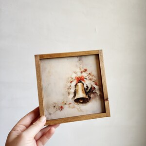 May include: A framed vintage Christmas print featuring a gold bell with red bows and holly leaves on a white background.
