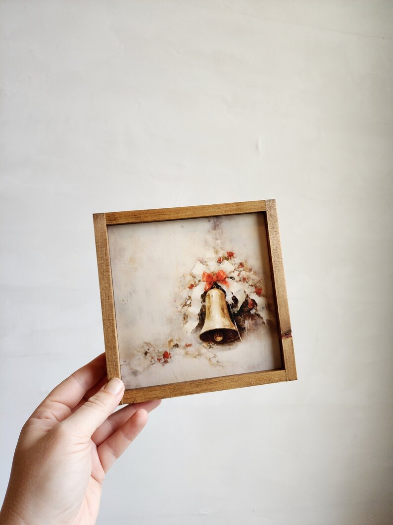 May include: A framed vintage Christmas print featuring a gold bell with red bows and holly leaves on a white background.
