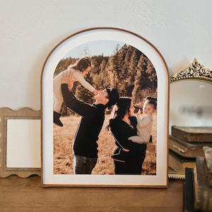 May include: A framed photograph of a family of four in a forest setting. The father is holding up a young girl, while the mother holds a baby girl. The family is smiling and looking at each other.