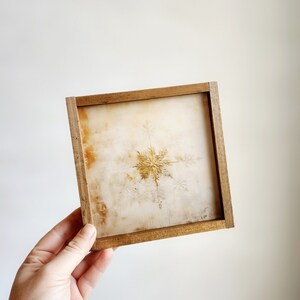 May include: A framed artwork featuring a gold snowflake on a white background.