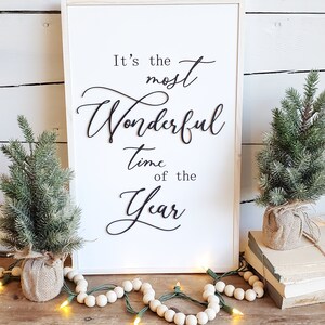 May include: A framed white sign with the text "It's the most Wonderful time of the Year" in black script. Two small evergreen trees in burlap pots flank the sign, with string lights and wooden beads.