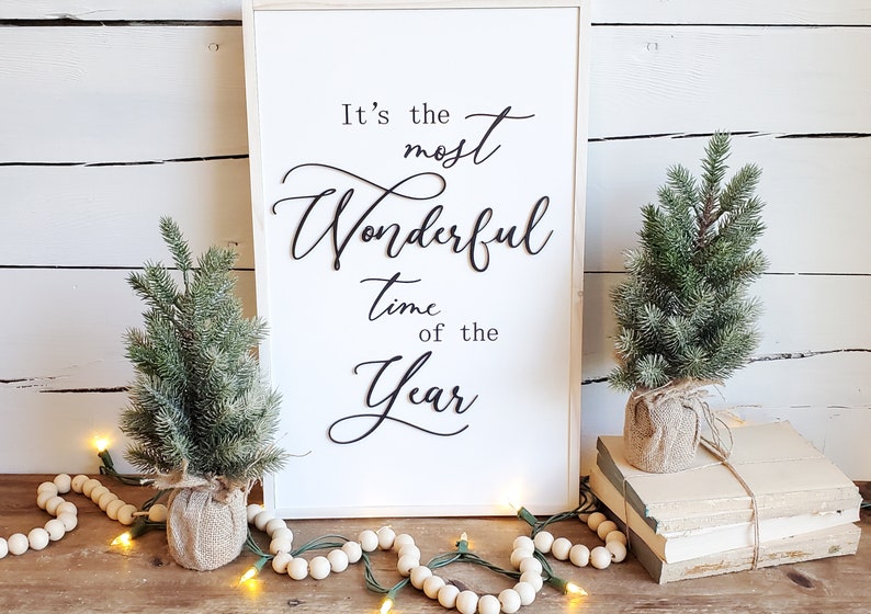 May include: A framed white sign with the text "It's the most Wonderful time of the Year" in black script. Two small evergreen trees in burlap pots flank the sign, with string lights and wooden beads.