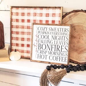May include: A wooden sign with the text "Cozy Sweaters, Pumpkin Everything, Cool Nights, Falling Leaves, Bonfires, Crisp Mornings, Hot Coffee" in black lettering. The sign is on a white shelf with a red and white checkered background and a wooden log slice behind it.