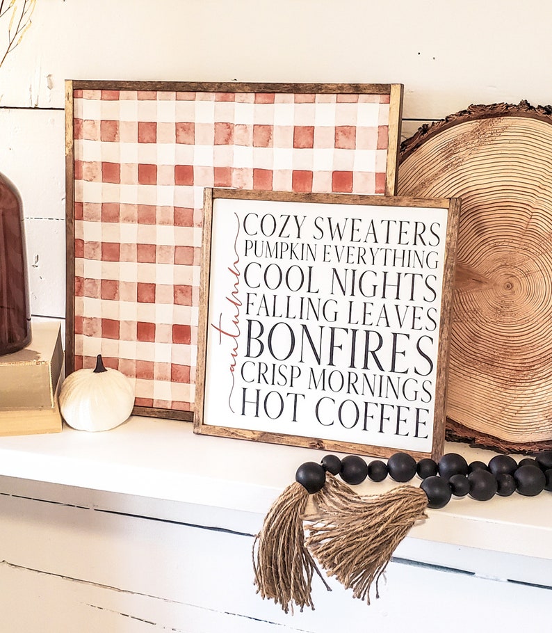 May include: A wooden sign with the text "Cozy Sweaters, Pumpkin Everything, Cool Nights, Falling Leaves, Bonfires, Crisp Mornings, Hot Coffee" in black lettering. The sign is on a white shelf with a red and white checkered background and a wooden log slice behind it.