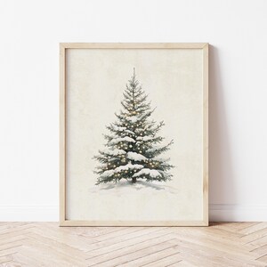 May include: A framed print of a snowy evergreen tree with white lights. The tree is centered on a cream background.