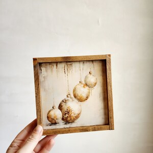 May include: A framed painting of four gold and white Christmas ornaments hanging from strings. The ornaments are painted in a watercolor style and have a rustic, vintage feel.
