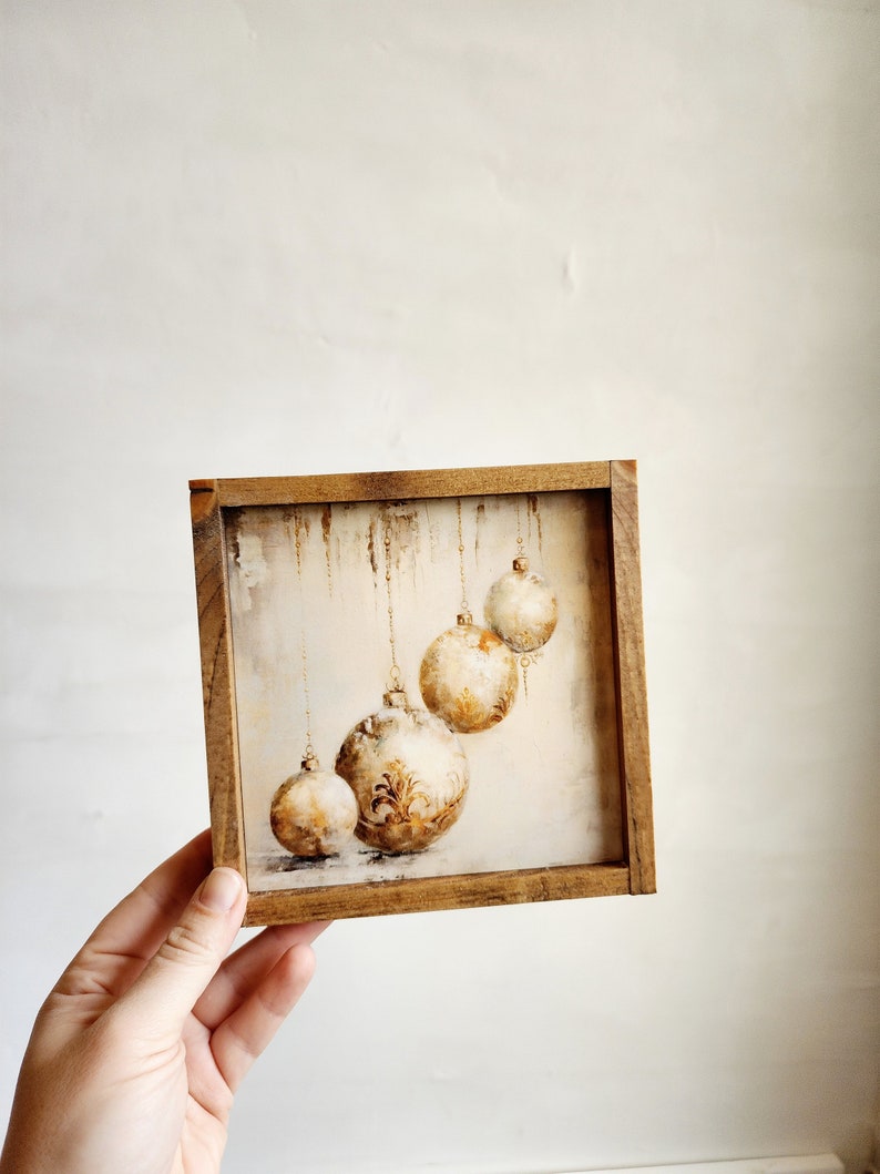 May include: A framed painting of four gold and white Christmas ornaments hanging from strings. The ornaments are painted in a watercolor style and have a rustic, vintage feel.