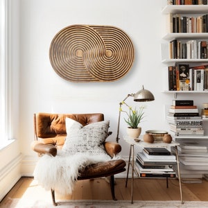 May include: A brown wooden wall art piece with a geometric design of concentric circles. The art is hanging on a white wall above a brown leather armchair with a white throw blanket.