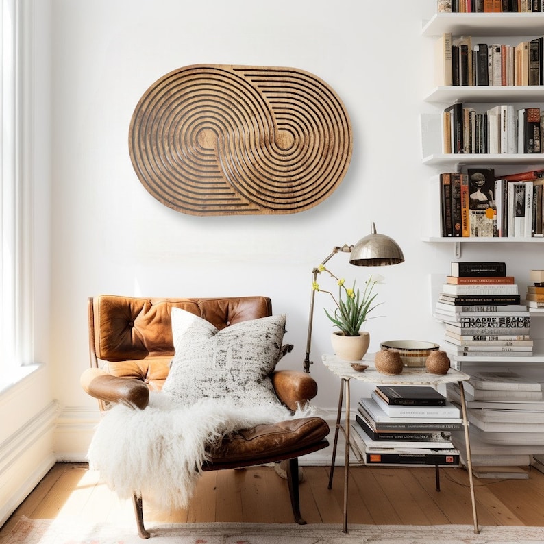 May include: A brown wooden wall art piece with a geometric design of concentric circles. The art is hanging on a white wall above a brown leather armchair with a white throw blanket.