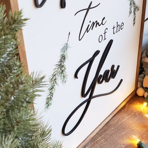 May include: Wooden sign with the words "Wonderful Time of the Year" in black script. The sign has a white background and a wooden frame. Decorative greenery and a string of lights are also visible.