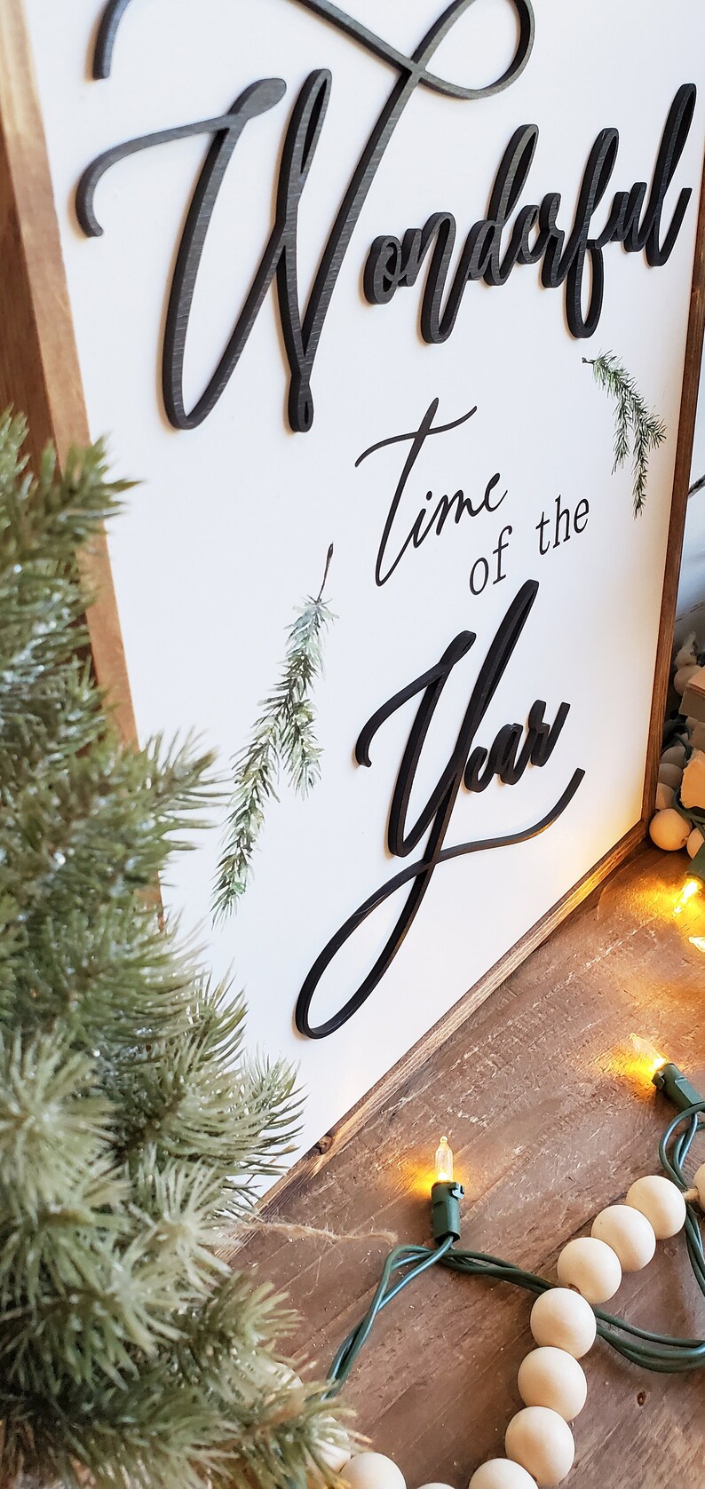 May include: Wooden sign with the words "Wonderful Time of the Year" in black script. The sign has a white background and a wooden frame. Decorative greenery and a string of lights are also visible.