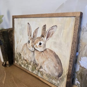 May include: A framed painting of two brown rabbits with white fur on their chests, sitting in a field of green grass. The painting has a light yellow background and is framed in a natural wood frame.
