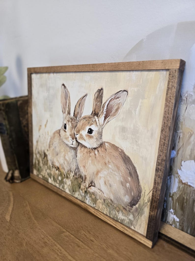 May include: A framed painting of two brown rabbits with white fur on their chests, sitting in a field of green grass. The painting has a light yellow background and is framed in a natural wood frame.