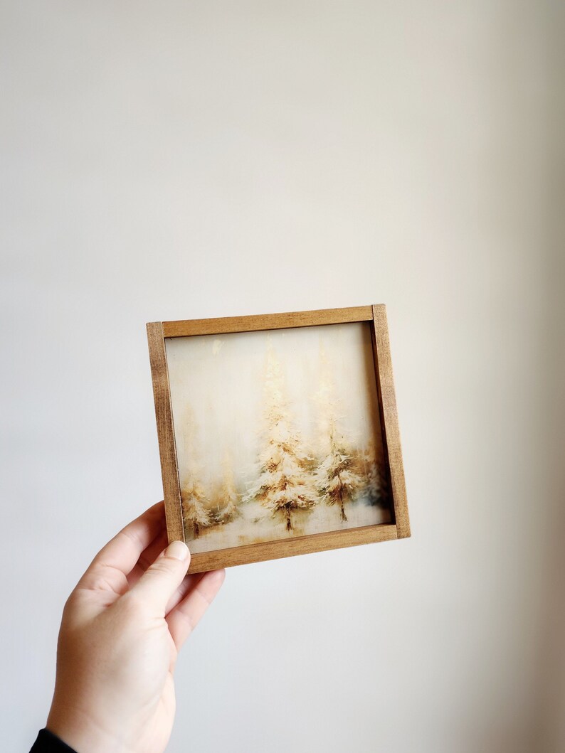 May include: A framed watercolor painting of a snowy forest scene with three trees in the center. The trees are painted in shades of brown, white, and gray, and the background is a soft, hazy blue.